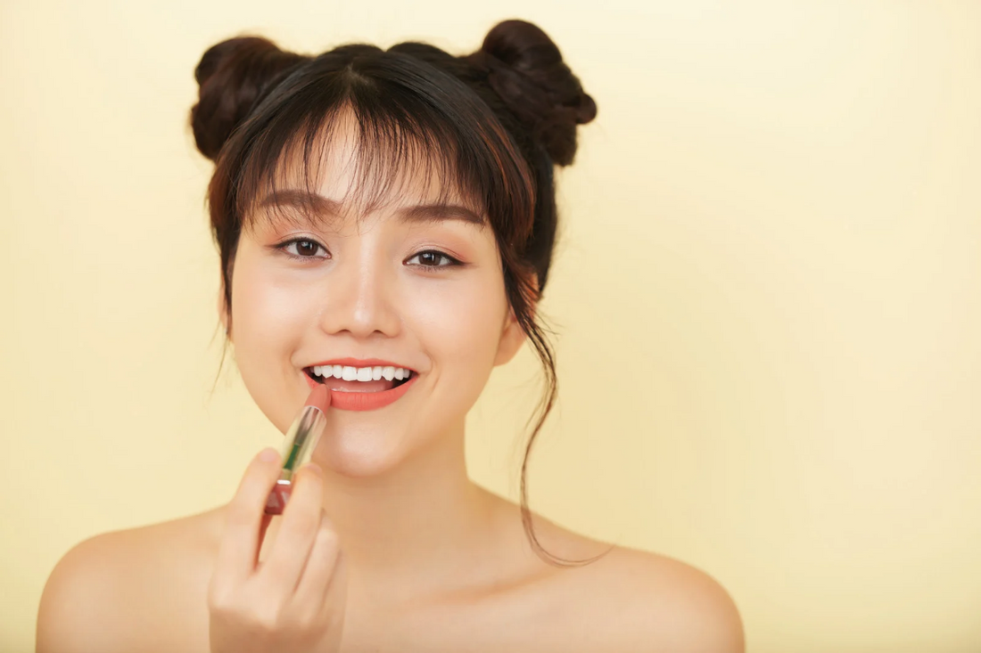 Woman applying makeup in natural light, wearing layered gold necklaces and simple stud earrings—capturing a stylish morning routine with everyday elegance.