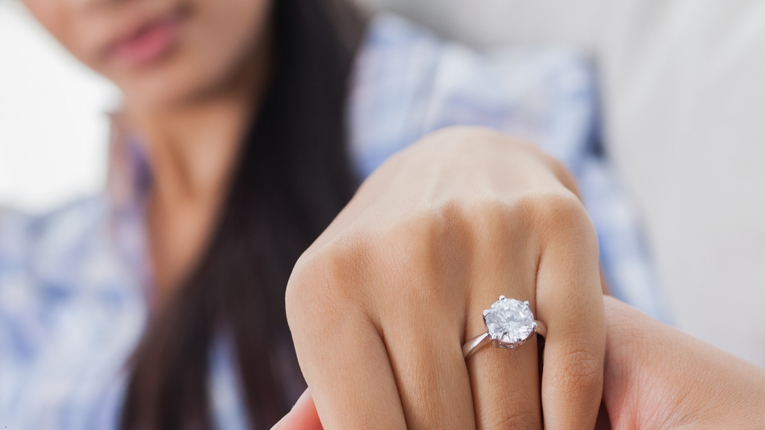 Close-up of a woman’s hand with a diamond engagement ring, focusing on the ring against a blurred background.